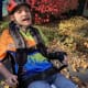 A man with cerebral palsy, wearing a helmet, colorful t-shirt, and jeans, smiles from his recumbent tricycle.
